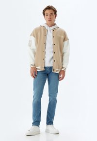 Chaqueta bloqueada en color beige y blanco con puños acanalados, cierre de botones y texto gráfico. Usada sobre una sudadera blanca y combinada con jeans azules.