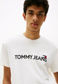 Camiseta de algodón blanca con un logo negro desgastado que dice "TOMMY JEANS" en el pecho y un pequeño parche de la marca en la manga izquierda.