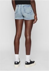 Šviesiai mėlyni džinsiniai šortai su sukabintais kraštais, turintys dvi nugarines kišenes, derinami su juodais sportiniais bateliais ir baltais kojinėmis. Lygus paviršius visame šortų plote.