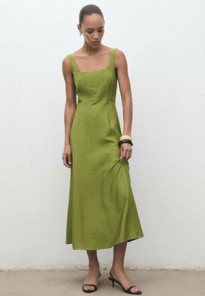 Grünes Kleid mit quadratischem Ausschnitt, ärmellosem Design und knöchellanger Rocklänge. Textil mit Struktur und dezenten Falten. Getragen mit schwarzen Sandalen.