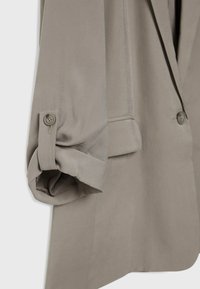 Manche d'un blazer beige roulée et maintenue par une patte boutonnée, montrant une poche avant partielle et une fermeture à un seul bouton sur le devant.