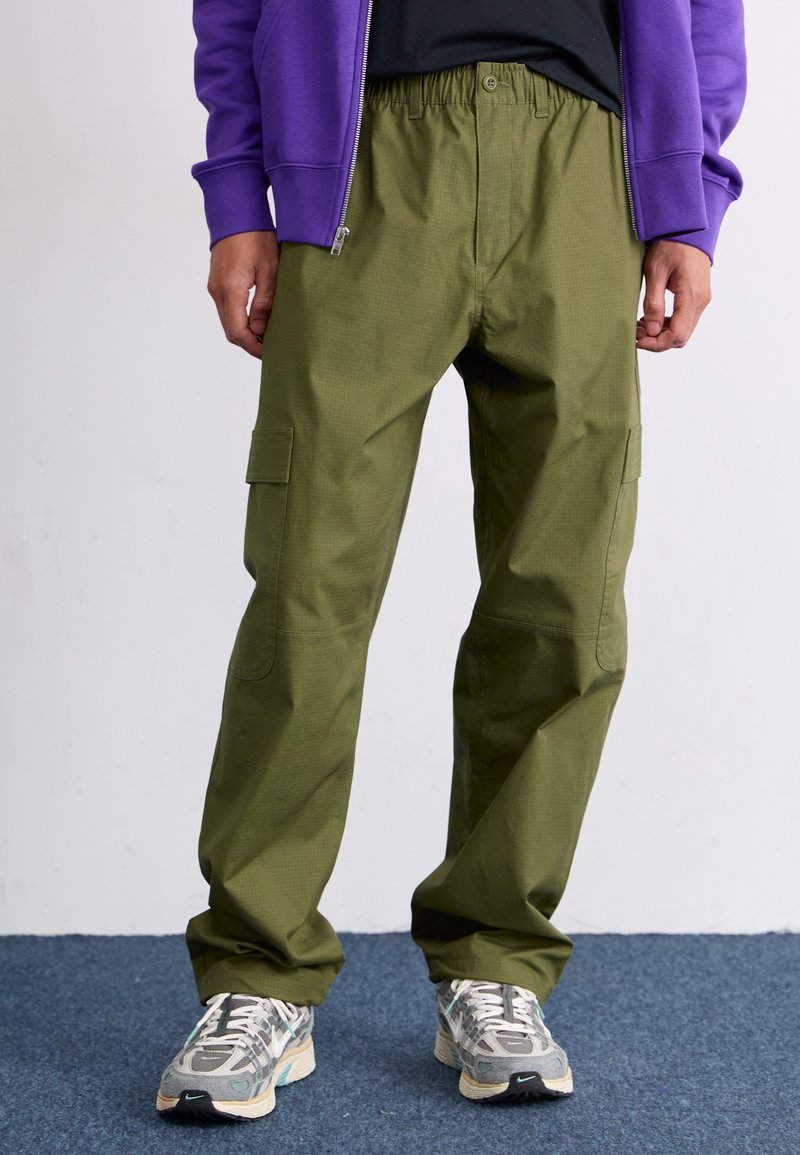 Pantalons cargo vert olive avec une taille élastique, deux poches latérales et une coupe ample, associés à des baskets grises et blanches à motifs.