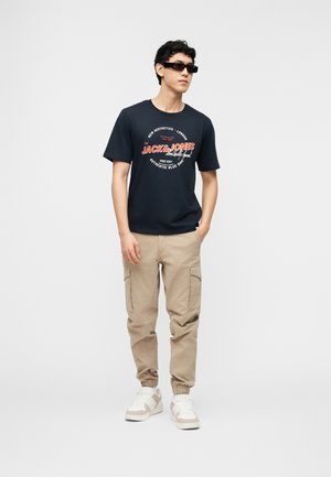 Mand iført sort T-shirt med grafisk print, beige cargobukser, hvide sneakers og rektangulære sorte solbriller mod en ensartet hvid baggrund.