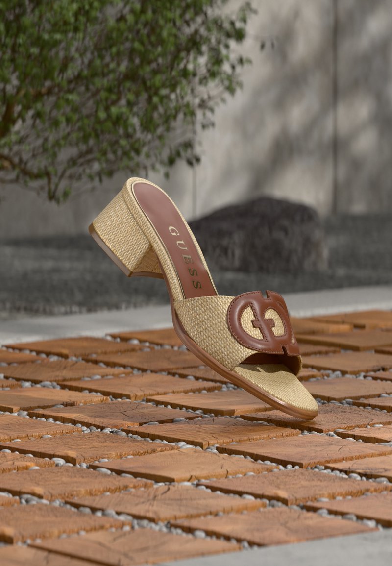 Μπεζ πλεγμένο σανδάλι με τακούνι και καφέ δερμάτινο λουράκι με κομμένο λογότυπο, τοποθετημένο πάνω σε επιφάνεια από τερακότα έξω, με φυτά στο φόντο.