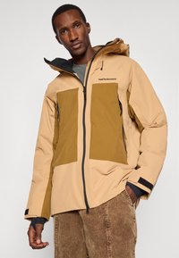 Άνδρας με μπεζ και καφέ φούτερ με κουκούλα, διαθέτει μπροστινό φερμουάρ και καφέ παντελόνι από καστόρι, στέκεται μπροστά από απλό φόντο.