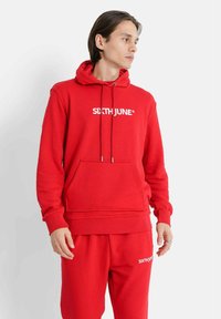 Röd hoodie i bomullsmix, med framficka i kängurustil, justerbar dragsko på huvan och vit logotyptext. Matchas med byxor i samma färg.