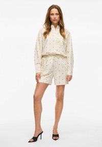 Beżowy, wzorzysty dwuczęściowy komplet z koszulą na guziki z kieszenią oraz szortami ze sznurkiem. Nosi się z czarnymi sandałami na obcasie.