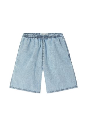 Lyshåret barn står iført en hvid langærmet skjorte med rød tekst, lyseblå shorts og brune sandaler mod en ensfarvet hvid baggrund.