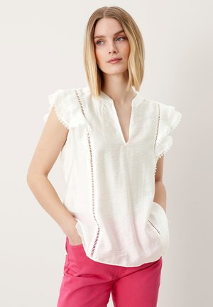 Blusa bianca leggera con maniche corte a balze, scollatura a V e dettagli in pizzo lungo le cuciture, abbinata a pantaloni rosa.