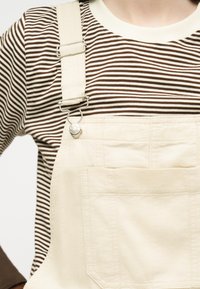 Person som har på seg en brun- og hvitstripete langermet skjorte under kremfargede kjeledresser med metallspenne og frontlomme.