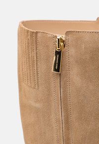 Sac à main en daim beige présentant une texture côtelée verticale et une fermeture éclair dorée avec un tire-zip logo. Intérieur lisse avec des détails de couture minimaux.