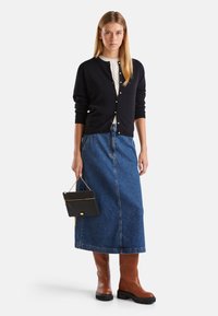 Námořnický kardigan s bílými knoflíky, krémový pletený top s boby, modrá džínová midi sukně, hnědé kozačky po kolena, držící černou kabelku se zlatým řetězem.