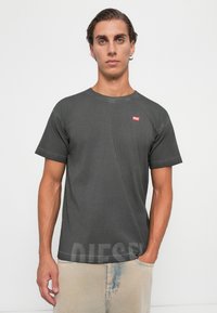 Jeune homme avec des cheveux gominés, portant un t-shirt noir Diesel délavé et un pantalon beige clair, se tenant devant un fond uni.