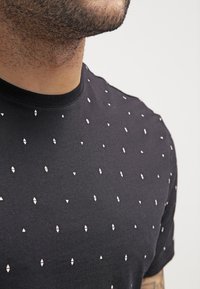 Czarny t-shirt z krótkim rękawem z teksturowanym wzorem małych białych trójkątów i owalnych kształtów, okrągłym dekoltem i bawełnianym materiałem.