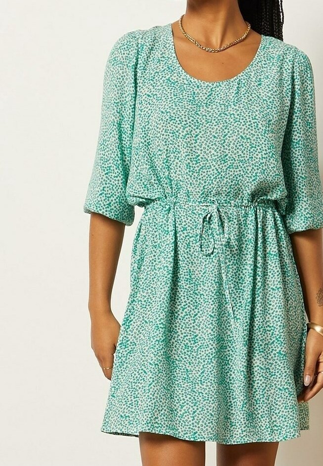 Femme portant une robe verte et blanche à motifs avec manches 3/4, une taille resserrée par un cordon et un collier chaîne doré.