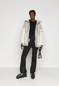 Μπουφάν σκι σε ανοιχτό γκρι και λευκό με φερμουάρ, κουκούλα και λεπτομέρειες. Συνδυασμένο με μαύρο παντελόνι και μαύρες μπότες σκι. Κρατώντας γυαλιά σκι.