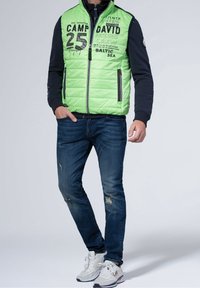 Zielona pikowana kamizelka z czarnym zamkiem błyskawicznym i nadrukowanym tekstem, w połączeniu z przetartymi niebieskimi dżinsami i białymi trampkami z szarymi akcentami.