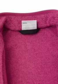 Fleece-Pullover in lebhaftem Pink mit einem strukturierten Strickkragen und einem Stoffetikett im Inneren, das liest: "Ich war hier." Weiches, gemütliches Material.