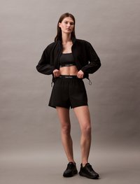 Mustvalge treeningriietus, mis koosneb lühikesest bomber-jakist, sportrinnahoidjast ja lühikestest pükstest. Pehme kangas, millega kaasnevad nöörid ja logo aktsendid. Mustad spordijalatsid.