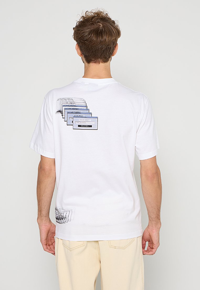 Mann trägt ein weißes T-Shirt mit blauen und schwarzen abstrakten rechteckigen Mustern auf dem Rücken, kombiniert mit hellbeigen Hosen.