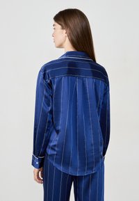 Marinblus i satin med vita vertikala ränder, lång ärm och rundad nederkant. Har en ryggsadel och kontrasterande knappdetalj på ärmsluten.