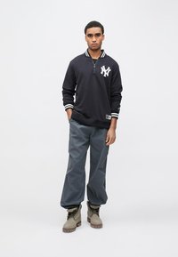 Juodas megztinis su baltu NY logotipu, kontrastingu dryžuotu apykakle, derinamas su laisvomis pilkomis kelnėmis ir smėlio spalvos bateliais su raišteliais.