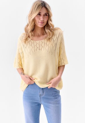 Pull en maille jaune clair à coupe ample, avec un motif texturé sur le haut et des manches courtes. Associé à un jean bleu.
