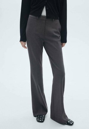 Osoba ubrana w ciemnoszare spodnie o prostych nogawkach, czarny kardigan, biały top i czarne, ćwiekowane buty z kwadratowym noskiem na jednolitym jasnym tle.