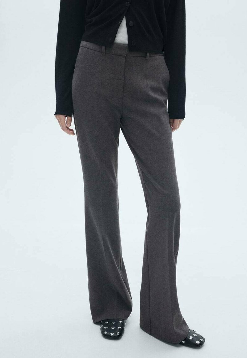 Personne portant un pantalon droit gris foncé, un cardigan noir, un haut blanc et des chaussures noires à bouts carrés cloutés sur un fond clair uni.