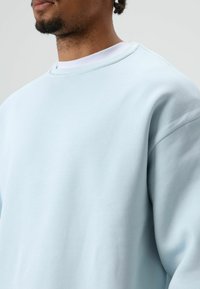 Sweatshirt bleu clair en tissu doux, avec un col rond et des épaules tombantes, présentant une texture lisse et des détails de couture minimaux.