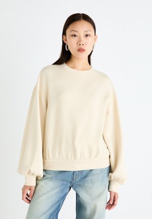 Beige oversized sweatshirt with long balloon sleeves, round neckline, and elastic hem. Worn with light blue jeans. Soft material texture.