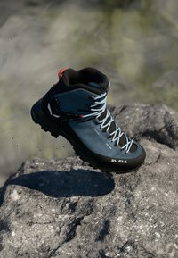 Scarpa da trekking blu con suola nera, dotata di lacci grigi, supporto alto alla caviglia e dettagli rossi. Superficie texturizzata e logo sul lato.