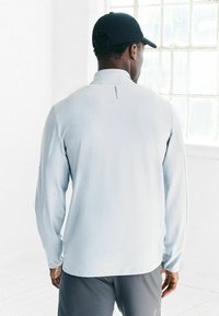 Vīrietis, ģērbies gaiši pelēkā garajās piedurknēs kreklā, tumši pelēkās biksēs un melnā cepurē, stāv telpā pie loga uz gaiša grīdas.