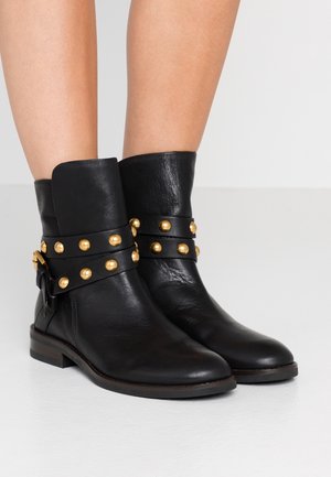 Botas negras de cuero hasta el tobillo con correas envolventes decoradas con tachuelas redondas doradas, usadas sobre piernas desnudas.