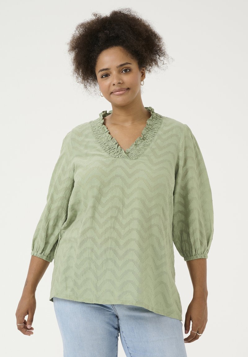 Blusa verde chiaro con un motivo a zigzag texture, scollatura a V con volant e maniche a tre quarti con polsini elasticizzati. Vestibilità casual.