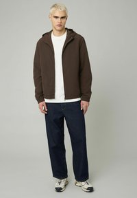 Veste légère zippée marron avec capuche, portée sur un t-shirt blanc, associée à un jean en denim sombre ample et des baskets multicolores.