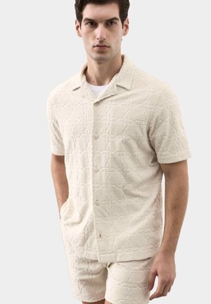 Hombre con camisa de manga corta con botón, beige y texturizada, y pantalones cortos a juego con patrón geométrico, de pie con las manos en los bolsillos.