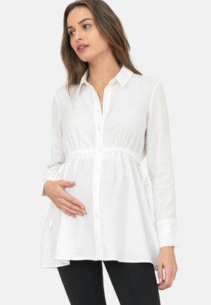 Mujer embarazada con cabello largo y castaño, vistiendo una blusa de maternidad blanca con botones y pantalones negros, sosteniendo suavemente su vientre.