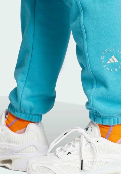Turkis sweatpants med manchetter ved anklerne, der har en logodetalje. Hvide sneakers med mesh-paneler og sokker med orange og lilla mønster.