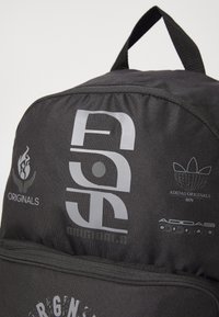 Sac à dos en tissu noir avec plusieurs logos argentés, une poignée supérieure et une poche zippée à l'avant avec une finition texturée.