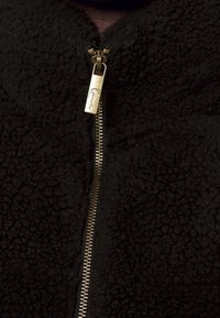 Giacca nera in pile con superficie strutturata, dotata di una zip dorata e una linguetta con un motivo. Colletto alto, cuciture minime.