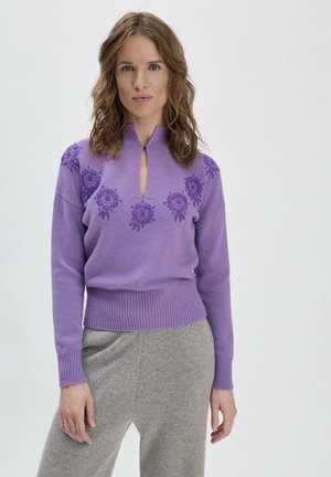 Lila gestrickter Pullover mit Stehkragen, ausgestattet mit floralem Stickerei auf der Brust und gerippten Bündchen, kombiniert mit grauen Hosen.