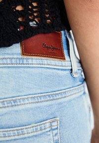 Világoskék farmer rövidnadrág barna bőr címkével. A címkén a "Pepe Jeans" felirat van domborítva. Részben látható fekete horgolt felső.