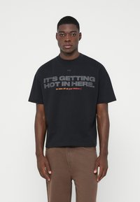 Νεαρός άνδρας που φοράει ένα μαύρο oversized T-shirt με το κείμενο "ΓΙΝΕΤΑΙ ΖΕΣΤΑ ΕΔΩ ΜΕΣΑ. οπότε βγάλε όλα σου τα ρούχα ;)" και καφέ παντελόνι.