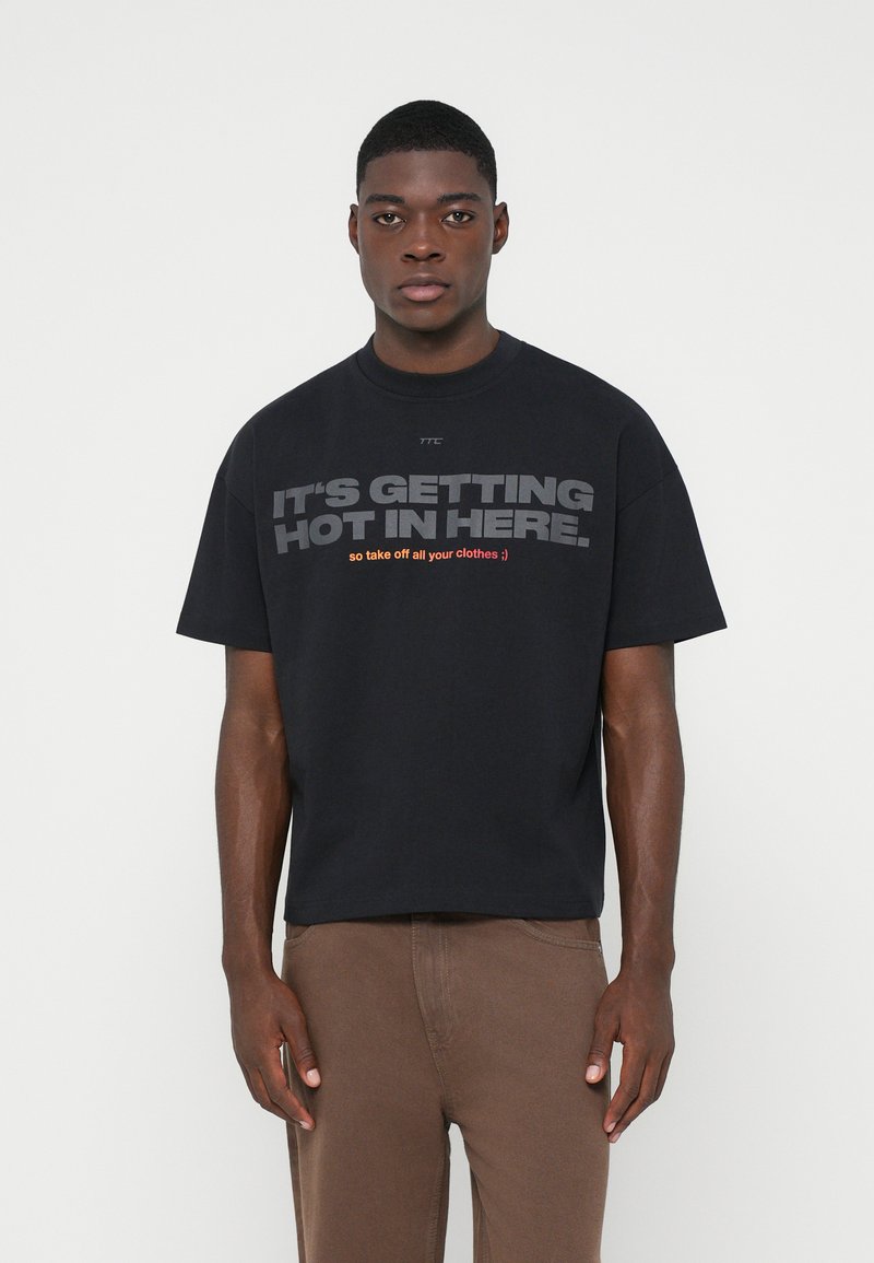 Νεαρός άνδρας που φοράει ένα μαύρο oversized T-shirt με το κείμενο "ΓΙΝΕΤΑΙ ΖΕΣΤΑ ΕΔΩ ΜΕΣΑ. οπότε βγάλε όλα σου τα ρούχα ;)" και καφέ παντελόνι.