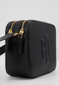 Černá kožená crossbody taška s texturovaným povrchem, se zlatými zipy a raženým logem na přední straně. Kompaktní obdélníkový tvar.