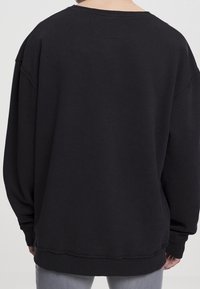 Personne portant un sweat-shirt noir uni à manches longues, vue de dos, associé à un pantalon gris clair.
