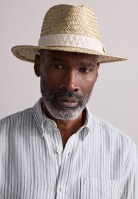 Chapeau fedora en paille à large bord avec un bandeau blanc, assorti à une chemise à boutons rayée bleu clair et blanc avec une poche poitrine.