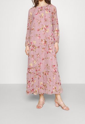 Femme portant une longue robe fleurie rose à manches longues et ourlet volanté, associée à des sandales à talons rose clair.