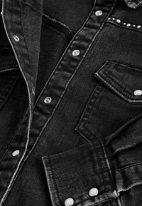 Gros plan d'une veste en denim noire montrant des boutons pression, une poche poitrine avec rabat et une rangée de petits clous décoratifs près du col.